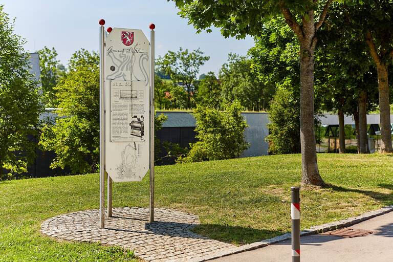 Eine Informationstafel steht auf einem gepflasterten Bereich in einem grünen Park, umgeben von Bäumen.