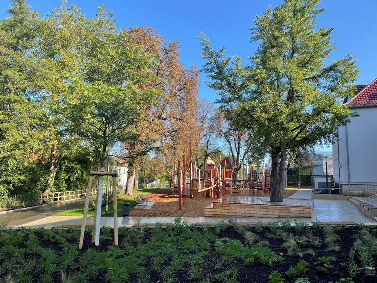 Das Bild zeigt einen sonnigen Spielplatz mit Klettergerüst umgeben von grünen Bäumen und einem gepflasterten Bereich.
