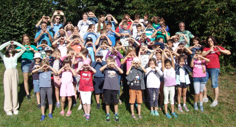 Eine große Gruppe von Kindern und Erwachsenen steht auf einer Wiese und formt mit den Händen ein Herzsymbol.