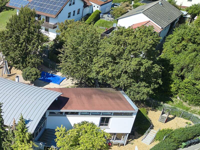Die Luftaufnahme zeigt eine Wohngegend mit mehreren Häusern, von denen eines Solarpaneele auf dem Dach hat, umgeben von Bäumen und Gärten.