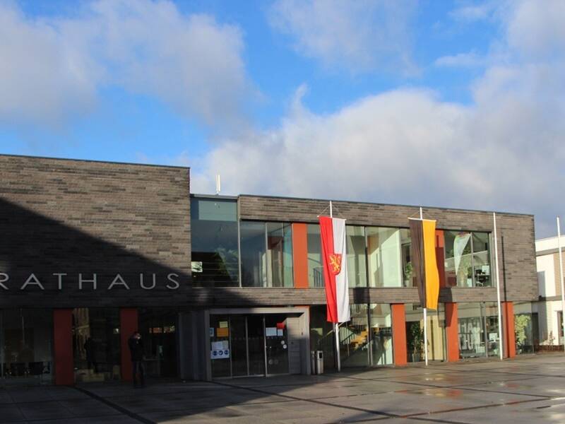 Das Bild zeigt ein modernes Rathausgebäude mit großen Glasfenstern und zwei Fahnen an der Fassade, beleuchtet von Sonnenlicht unter einem blauen Himmel mit Wolken.