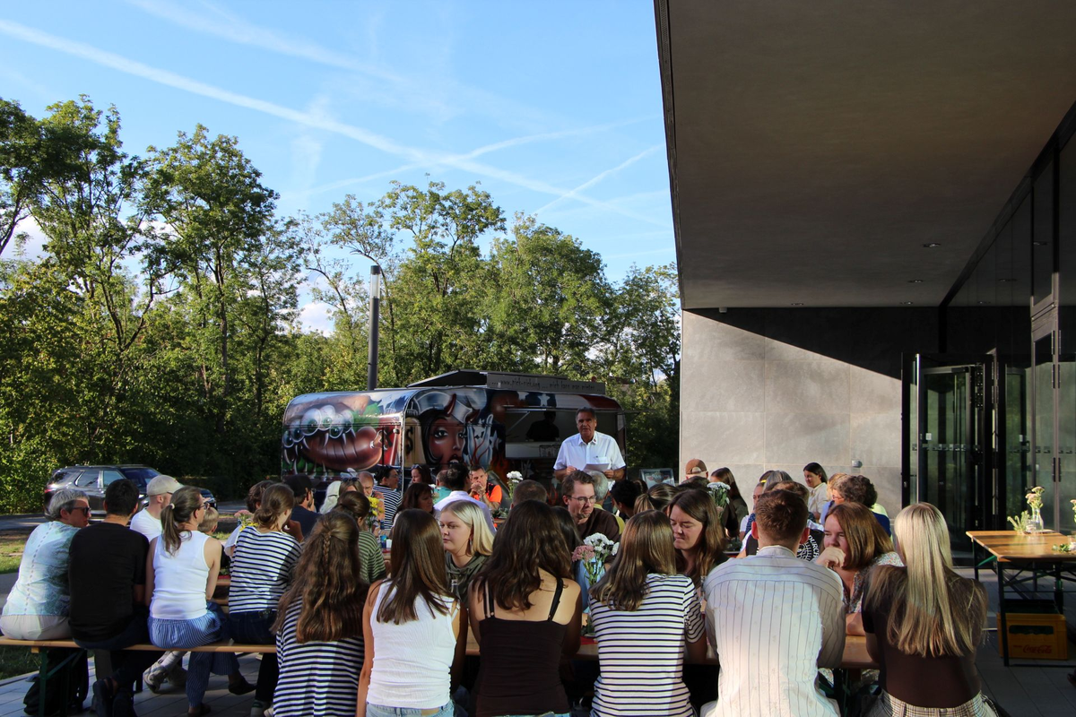 Eine Gruppe von Menschen sitzt an langen Biertischen im Freien und unterhält sich, während im Hintergrund ein bunt bemalter Foodtruck zu sehen ist.