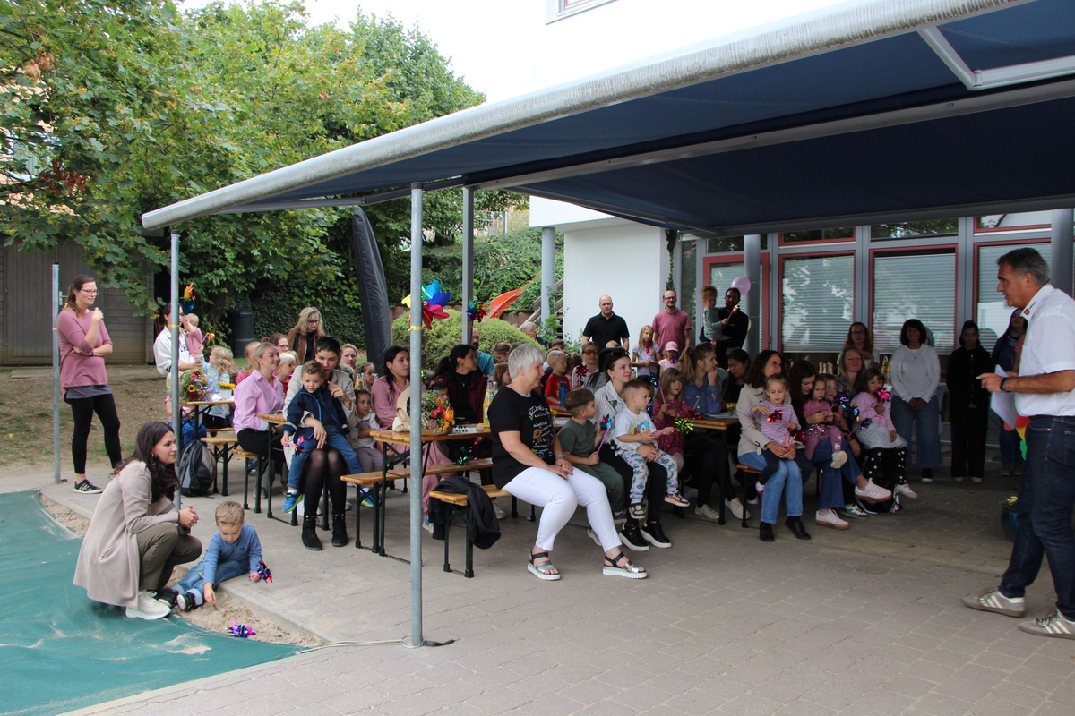 Eine Gruppe von Erwachsenen und Kindern sitzt und steht unter einem überdachten Bereich, während ein Mann vorne zu ihnen spricht.