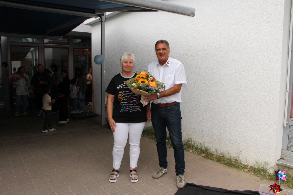 Eine Frau und ein Mann stehen draußen vor einem Gebäude, wobei der Mann der Frau einen Blumenstrauß überreicht.