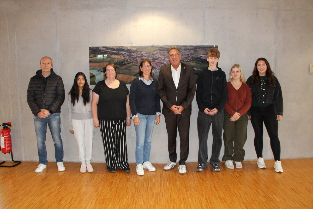 Eine Gruppe von acht Menschen, bestehend aus Männern und Frauen, steht in einer Reihe vor einer Wand mit einem Bild.