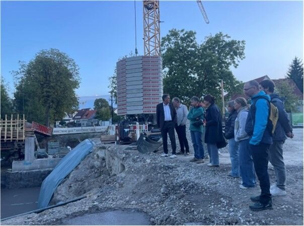 Eine Gruppe von Menschen steht auf einer Baustelle neben einem Kran und einer großen Konstruktion von gestapelten Elementen, umgeben von Bäumen und Gebäuden im Hintergrund.
