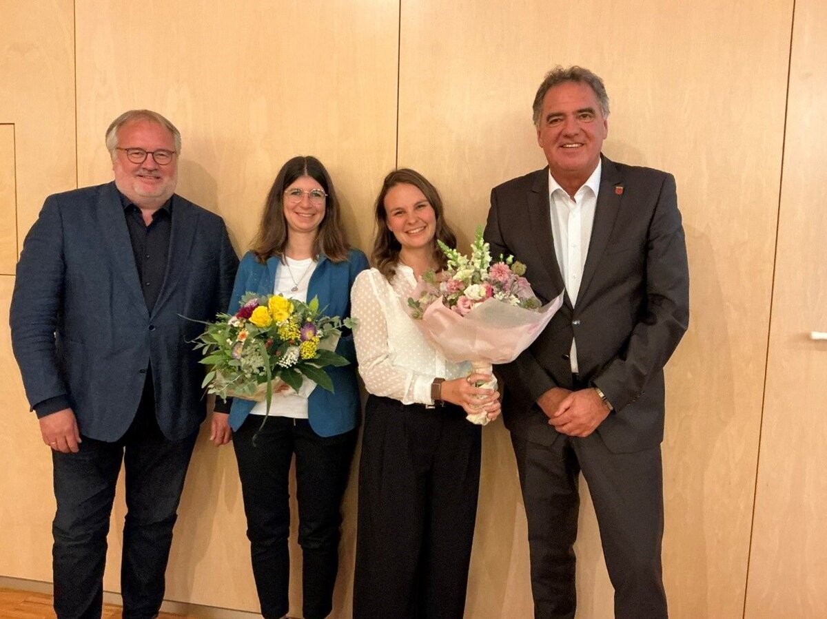 Vier Personen stehen lächelnd nebeneinander, zwei davon halten Blumensträuße in den Händen.