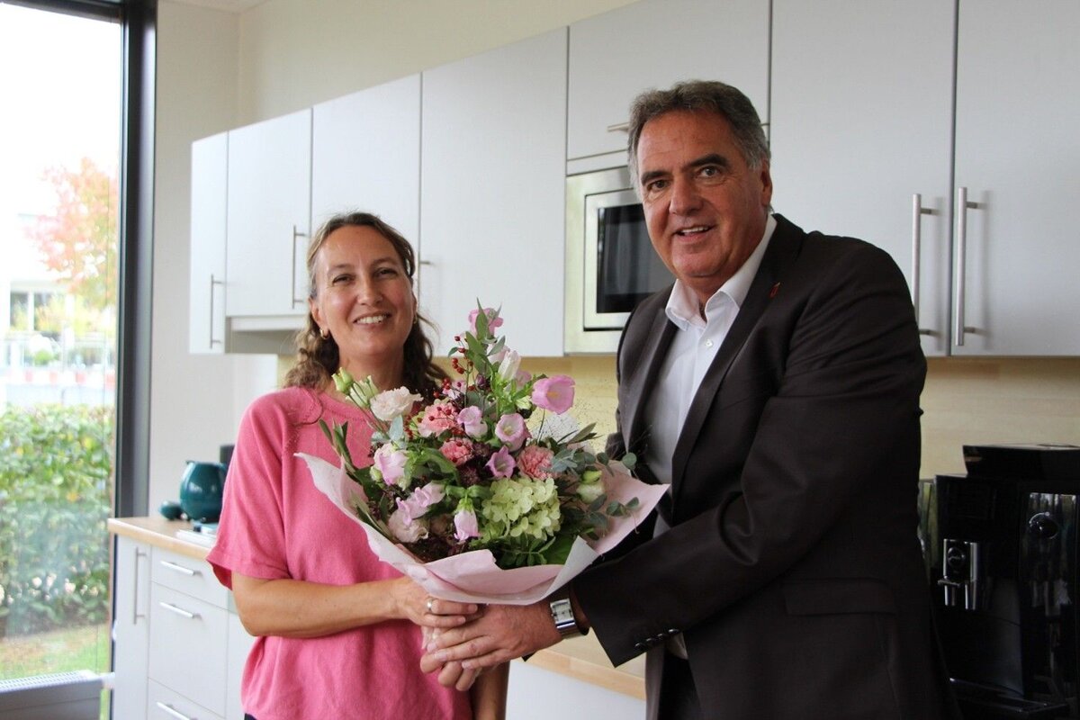 Ein Mann im Anzug überreicht einer lächelnden Frau in einem rosa Oberteil einen Blumenstrauß in einer modernen Küche.
