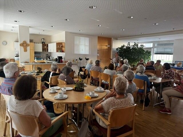 Eine Gruppe älterer Menschen sitzt in einem Gemeinschaftsraum, der wie ein Café gestaltet ist, und schaut auf eine Präsentation an der Wand.