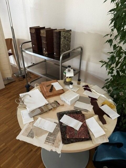 Ein Tisch mit verschiedenen Buchbinder-Utensilien, darunter Stoffproben und Werkzeuge, steht neben einem Metallschrank mit großen, alten Büchern.