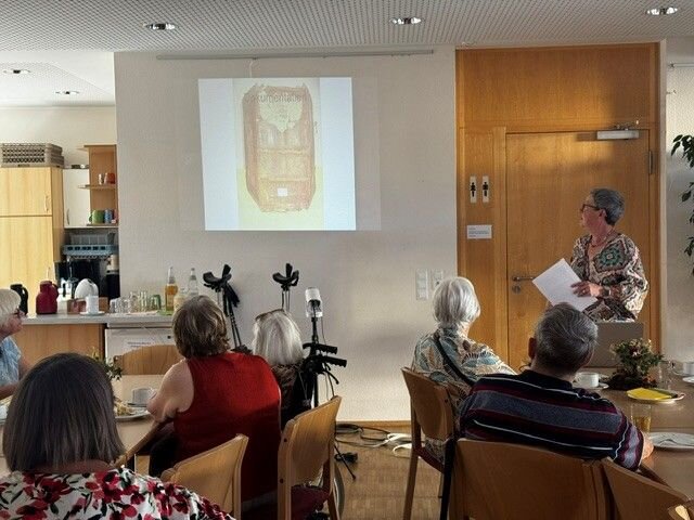 Eine Person hält einen Vortrag vor einer Gruppe älterer Menschen in einem Raum mit einem projizierten Bild an der Wand.