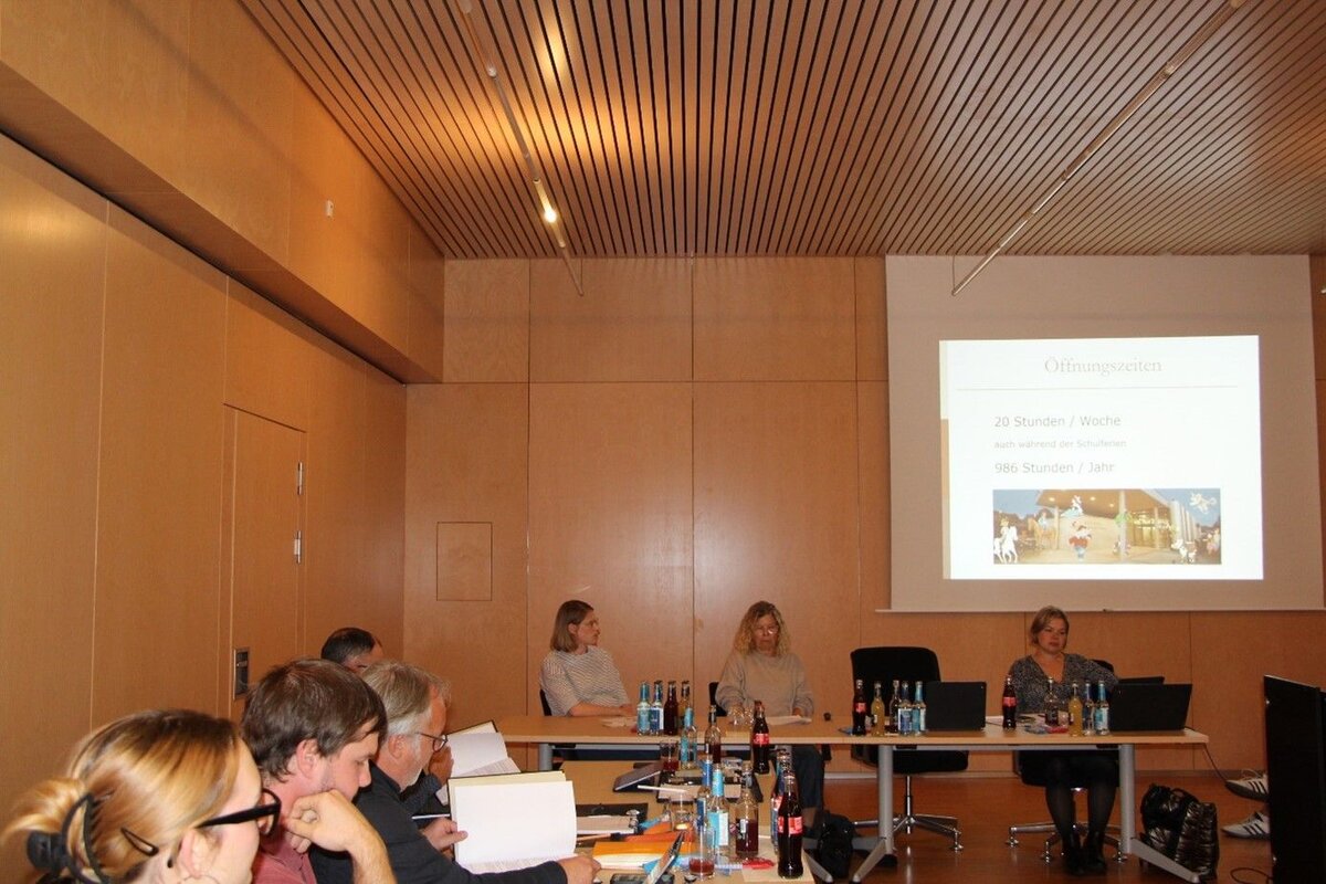 Eine Gruppe von Menschen sitzt in einem Konferenzraum an einem langen Tisch, während eine Präsentation mit dem Titel "Öffnungszeiten" auf eine Leinwand projiziert wird.