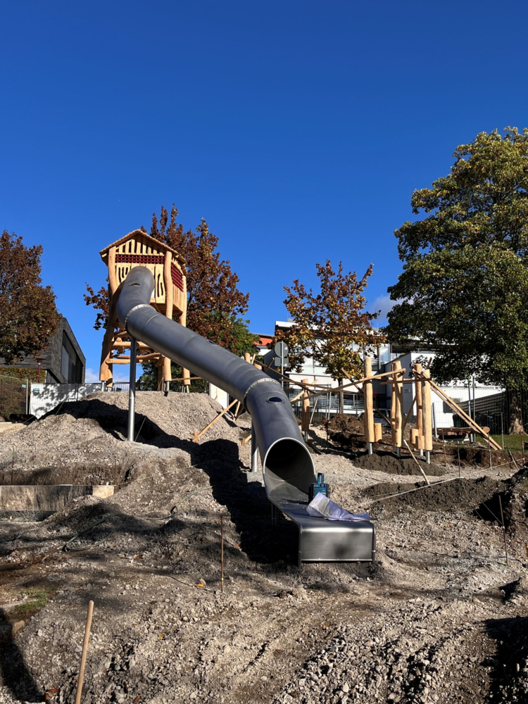 Eine große Rutsche aus Metall führt von einem erhöhten hölzernen Spielturm hinunter auf einen mit Erde bedeckten Spielplatz unter einem klaren blauen Himmel.