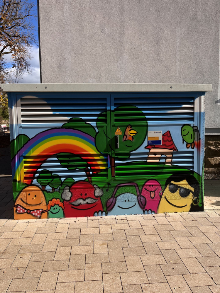 Ein Verteilerkasten ist mit buntem Graffiti bemalt, das fröhliche Gesichter, einen Regenbogen und Landschaftselemente zeigt.