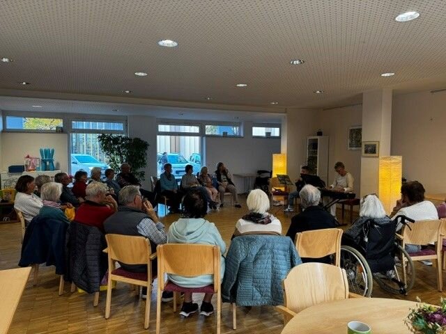 Eine Gruppe älterer Menschen sitzt in einem Raum in einem Stuhlkreis und hört einem Musiker am Keyboard zu.