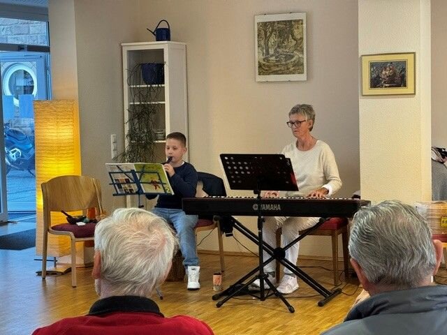 Ein Junge spielt Flöte in ein Mikrofon, während eine Frau Klavier spielt; sie sitzen nebeneinander in einem Raum mit Publikum.