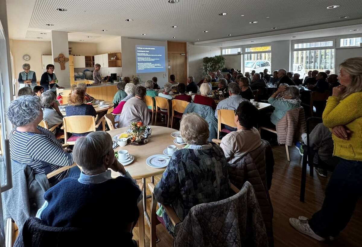 Eine Gruppe älterer Menschen sitzt in einem hellen Raum, während vorne eine Präsentation gezeigt wird.