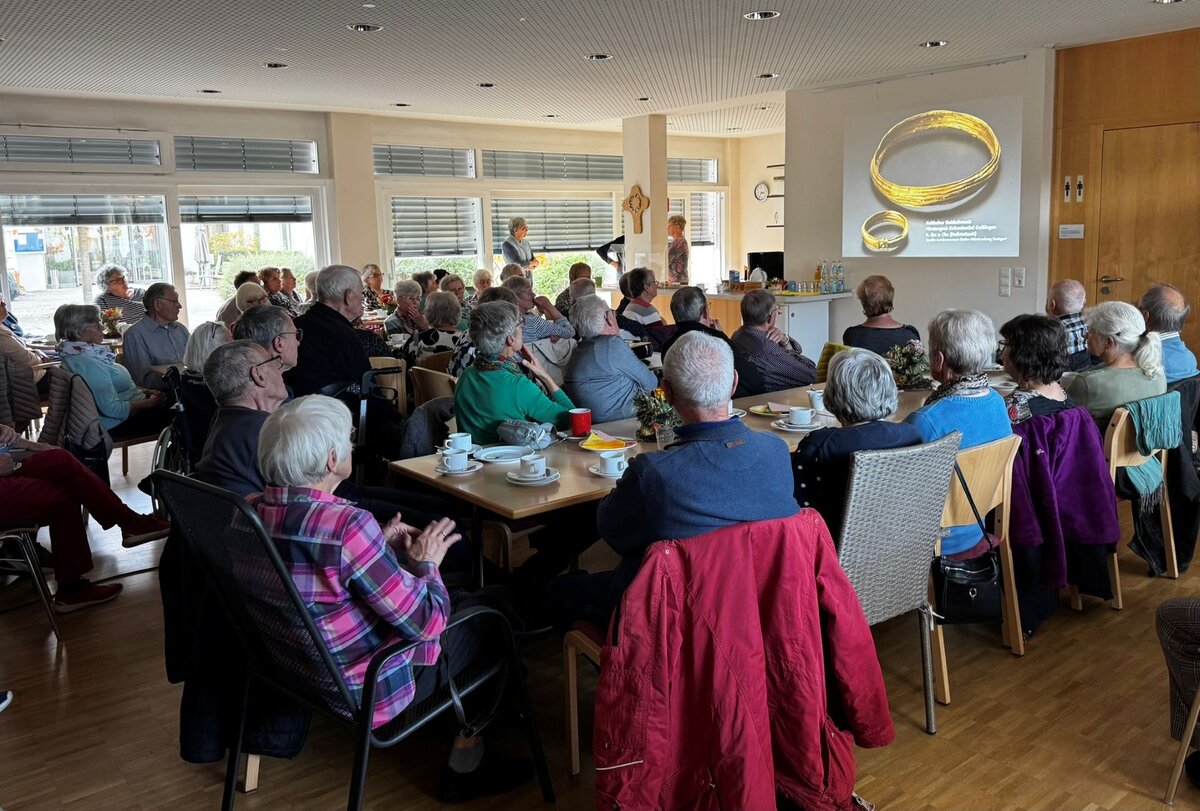 Eine Gruppe älterer Menschen sitzt in einem Raum und schaut sich eine Präsentation über antiken Schmuck auf einer Leinwand an.