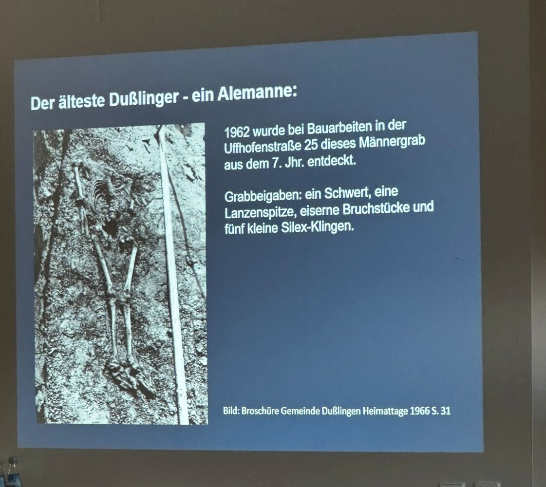 Die Präsentation zeigt ein altes Foto eines im Jahr 1962 entdeckten Männergrabs aus dem 7. Jahrhundert, zusammen mit einem Text, der die Grabbeigaben beschreibt.