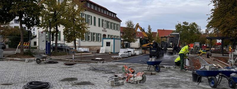Vor einem großen, weißen Gebäude mit der Aufschrift "Rathaus" arbeiten mehrere Personen auf einer Baustelle, umgeben von Baumaschinen und Baumaterialien.
